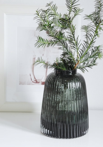 Wazon szklany ryflowany z ciemnego szkła z zielonymi gałązkami na białym blacie