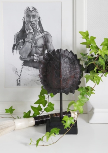 Dekoracyjna skorupa żółwia w kolorze ciemnobrązowym na czarnym, metalowym stojaku. Obok biały wazon z bluszczem i biało-brązowy pędzel w stylu etno