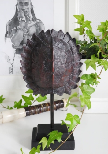 Egzotyczna figurka skorupa żółwia na postumencie stojąca na białym blacie. W tle oprawione czarno-białe zdjęcie etniczne i zielone liście bluszczu