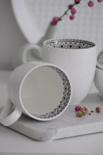 Zbliżenie na biały kubek porcelanowy: wyraźna faktura rombów na zewnętrznej ściance i kontrastowy, czarny wzór geometryczny widoczny w środku naczynia.