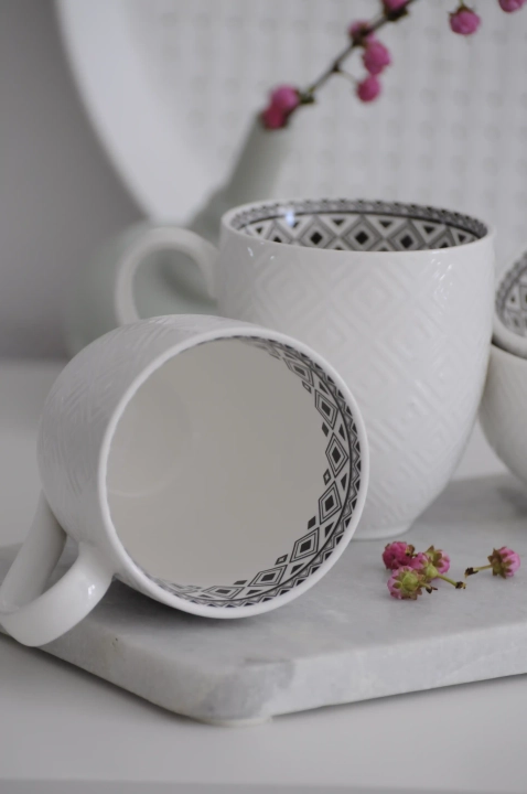 Zbliżenie na biały kubek porcelanowy: wyraźna faktura rombów na zewnętrznej ściance i kontrastowy, czarny wzór geometryczny widoczny w środku naczynia.