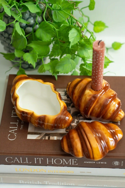 Otwarty ceramiczny pojemnik w kształcie rogalika, jego pokrywka oraz świecznik ze świecą na książkach w aranżacji domowej